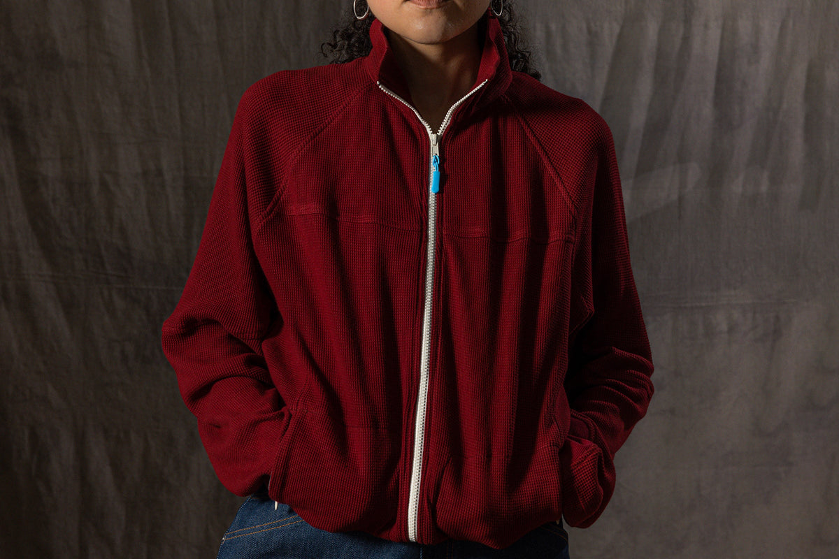 A person wearing a deep red fleece zip-up jacket with a white zipper, blue jeans, and a red and blue patterned knit hat, standing against a dark gray fabric backdrop.