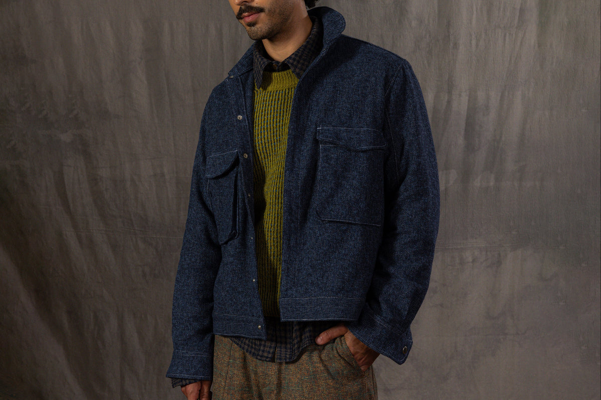 A man wearing a blue patterned knit beanie, a dark blue textured jacket over a green knit sweater, a dark plaid shirt, and brown patterned trousers, standing against a gray fabric backdrop.